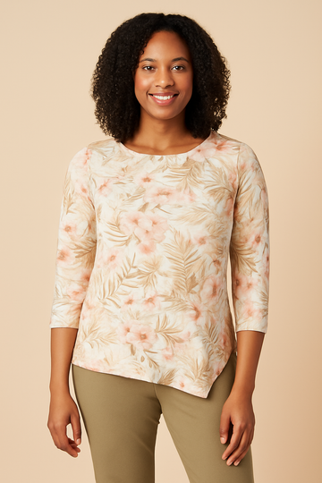 Front view of an African American woman with shoulder-length curly hair, smiling warmly while wearing a Comfort Code Baby French Terry Asymmetric Top in a soft floral print with blush and coral hues. She is paired with olive-green pants and stands against a soft beige background, highlighting the top’s asymmetric hem and delicate floral patterns for a fresh, elegant, and natural look.