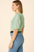 Side view of a young woman wearing a Democracy Embroidered Puff-Sleeve Top in soft green, paired with light blue high-waisted denim jeans and a black belt. The top features delicate eyelet embroidery on the puff sleeves. She accessorizes with a gold watch, a thin gold chain necklace, and gold hoop earrings, creating a casual yet elegant look.