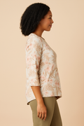 Side view of an African American woman with shoulder-length curly hair, smiling warmly while wearing a Comfort Code Baby French Terry Asymmetric Top in a soft floral print with blush and coral hues. She is paired with olive-green pants and stands against a soft beige background, highlighting the top’s asymmetric hem and delicate floral patterns for a fresh, elegant, and natural look.