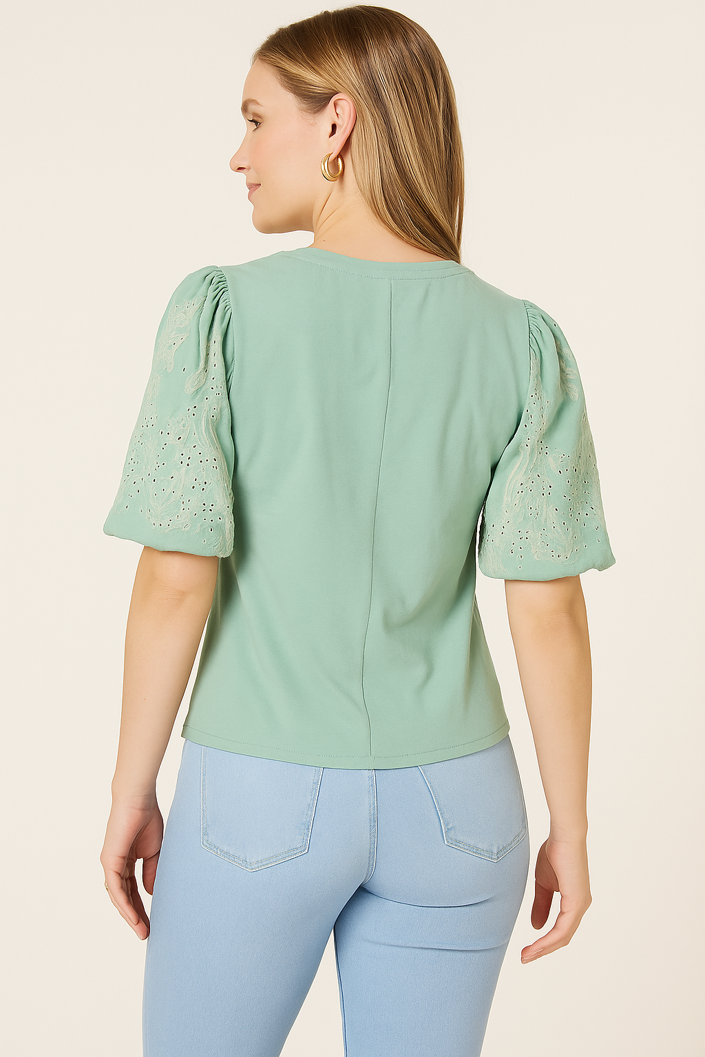 A young woman is shown from behind wearing a Democracy Embroidered Puff-Sleeve Top in sage green, paired with light blue high-waisted jeans. The top features intricate floral embroidery on the puff sleeves, while her straight, light blonde hair cascades down her back. Gold hoop earrings complete the look, all captured against a soft beige backdrop.