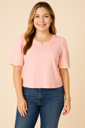 A young woman with fair skin smiles while wearing a Democracy Embroidered Puff-Sleeve Top in pastel pink, styled with classic blue denim jeans. The top features delicate eyelet embroidery on the puff sleeves, creating a soft and casual look. Her light brown wavy hair, subtle gold hoop earrings, and a natural makeup style complement the soft beige background.