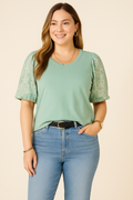A young woman with fair skin models a Democracy Embroidered Puff-Sleeve Top in sage green, styled with trendy light blue high-waisted jeans. The top features delicate lace embroidery on the puff sleeves, paired with a black leather belt, gold hoop earrings, a gold chain necklace, and a classic wristwatch, creating a stylish and polished look.