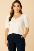 A young woman smiles while wearing a Democracy Embroidered Puff-Sleeve Top in white, paired with sleek navy blue tailored trousers. The top features delicate eyelet embroidery on the puff sleeves, giving it a sophisticated look. She accessorizes with gold hoop earrings, a delicate gold chain necklace, and a classic gold watch, creating a polished, semi-formal outfit against a soft beige background.