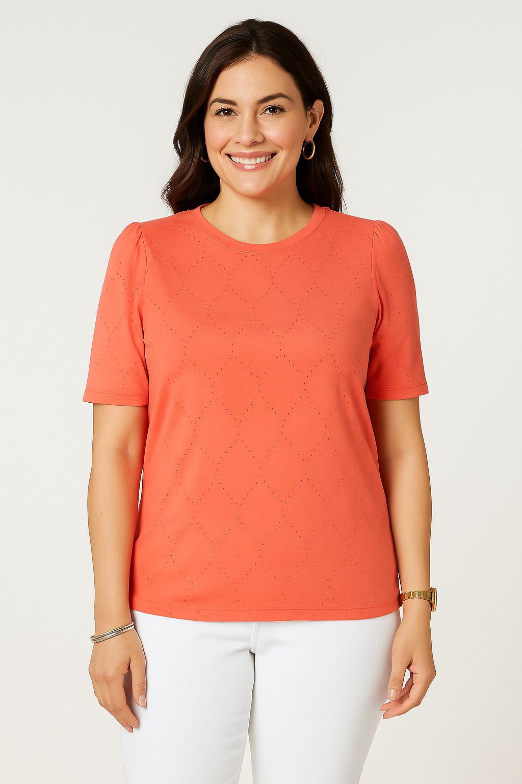 A woman smiles while wearing a Jaclyn Smith Eyelet Knit Puff Sleeve Tee in vibrant coral, paired with white pants. The soft eyelet pattern adds a delicate texture to the T-shirt, complemented by simple gold jewelry.