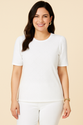 A woman with medium skin tone wears a Jaclyn Smith Eyelet Knit Puff Sleeve Tee in white, styled with matching white pants. She smiles confidently, accessorized with gold hoop earrings and a watch, set against a beige background.