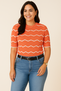 Jaclyn Smith Short-Sleeve Chevron Knit Sweater in bright coral with white chevron patterns, styled in a trendy look with light blue high-waisted jeans, a black leather belt, gold hoop earrings, and a wristwatch.

